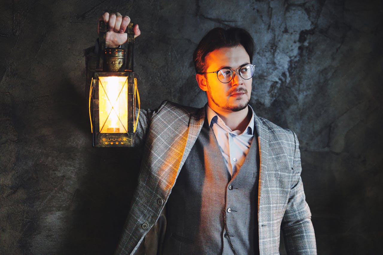 A man in a checkered suit holds a lantern casting warm light against a dark textured wall.